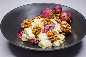Black dish. Large walnuts. Salad with chicken fillet and pink grapes. Dark background. White table. Concept for a tasty and healthy meal.