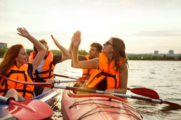 Happy young caucasian group of friends kayaking on river with sunset in the backgrounds. Having fun in leisure activity. Happy male and female model laughting on the kayak. Sport, relations concept. - obrazy, fototapety, plakaty