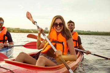 Happy young caucasian group of friends kayaking on river with sunset in the backgrounds. Having fun in leisure activity. Happy male and female model laughting on the kayak. Sport, relations concept.