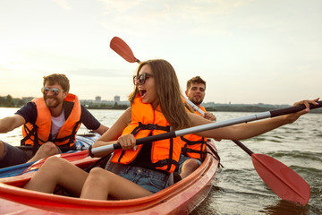Obraz premium Happy young caucasian group of friends kayaking on river with sunset in the backgrounds. Having fun in leisure activity. Happy male and female model laughting on the kayak. Sport, relations concept.