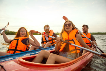 Happy young caucasian group of friends kayaking on river with sunset in the backgrounds. Having fun in leisure activity. Happy male and female model laughting on the kayak. Sport, relations concept.