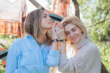 Portrait of two beautiful women brunette and blond holding their hands with same bracelets next to the tree in late summer. Beautiful girls standing next to each other.