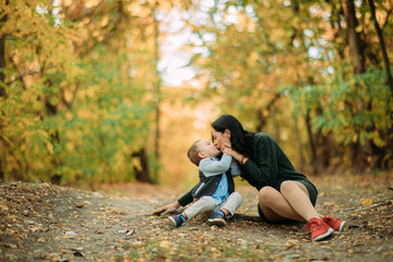 A mother sits on a pathway and kisses her son in autumn forest.