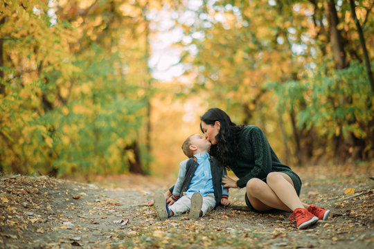 A mother sits on a pathway and kisses her son in autumn forest.