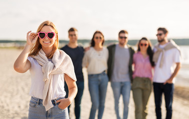 friendship, valentine's day and people concept - happy woman in heart-shaped sunglasses with group of friends on beach in summer