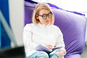 Young attractive smiling woman makes some notes in notepad at library and sitting on violet couch.