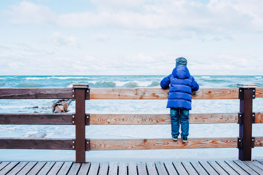 A lonely kid in blue jacket and jeans at the beach looking at the stormy sea staying on a fence. Winter or late autumn season