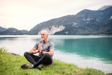 Naklejka na ściany i meble A senior man pensioner sitting by lake in nature, doing yoga exercise.