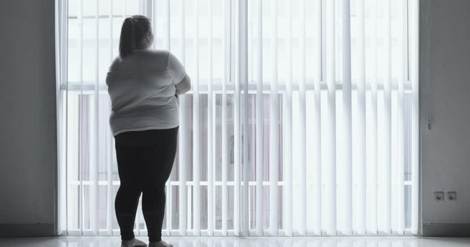 Silhouette of moody overweight woman standing alone near the window at home. Shot in 4k resolution