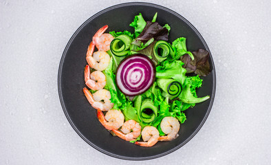 Salad with cabbage leaves and golden shrimps. Black dish. Dark background. White table. Concept for a tasty and healthy meal. Crimean onion. Green cucumber and arugula. Top view. Flat lay.