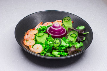 White table. Concept for a tasty and healthy meal. Green cucumber and arugula. Salad with cabbage leaves and golden shrimps. Black dish. Dark background. Crimean onion.