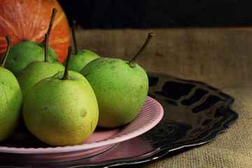 Green organic pears and one small red apple, different not like others, on delicate pink plate on silver metal tray, picturesque still life on coarse retro cloth canvas. Dark atmospheric fall harvest