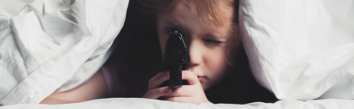 panoramic shot of scared child holding gun while hiding under blanket