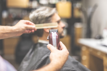 Professional barber holding comb and clipper, client sitting in armchair in the background.