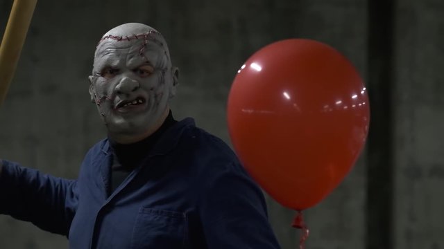Monster from movie froze with his hand raised, holds red ball, baseball bat, looks directly at camera, prepares for attack, scary man, basement, dark parking lot, dangerous place