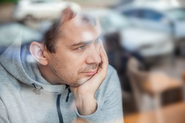 Sad pensive handsome man leaning head on hand in cafe, looking through a window with reflections.