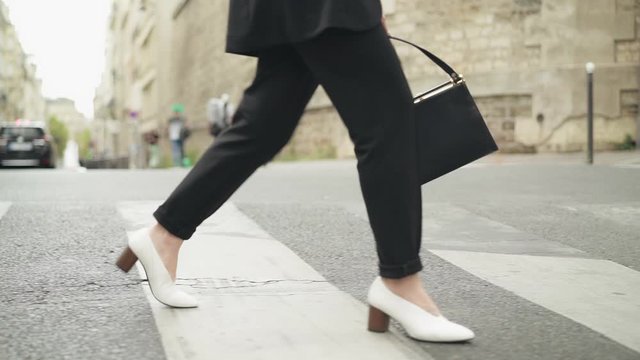 Slow motion of woman legs in black casual trousers in white boots walking on the road on zebra crossing, holding a small black bag