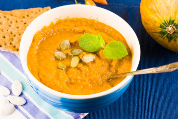 Pumpkin soup in a bowl is served with herbs, pumpkin seeds and rye bread. Vegan soup. Blue background.