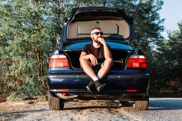 Cute bearded man with sunglasses sitting in the trunk of his car.