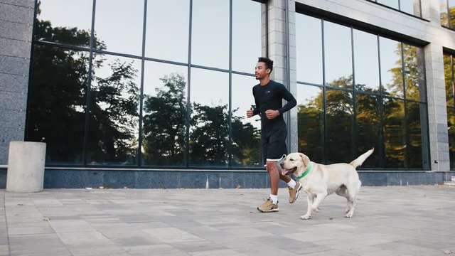 Young black man running with his white labrador dog in buseness city center during beautiful fall morning, slow motion