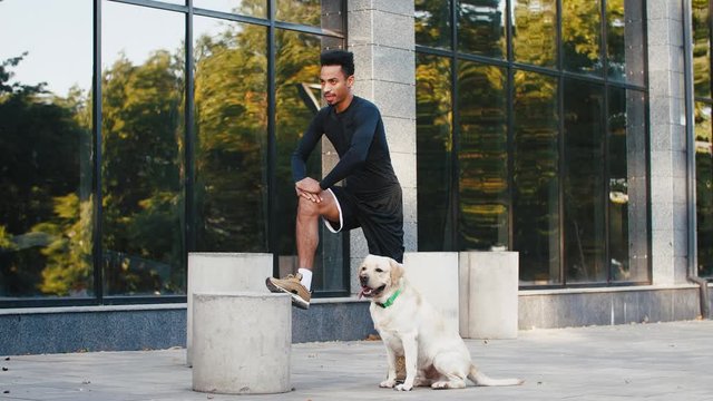 Young black man stretching after his training in city center with his white labrador dog beside him in the morning, slow motion