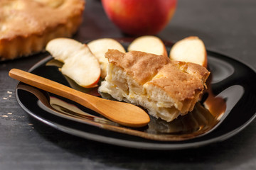 Slice Charlotte apple pie on a black plate with a wooden spoon on a black wooden table.Close up.