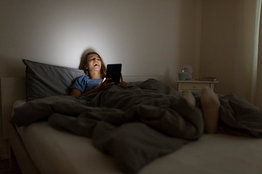 Laughing young woman lying in bed at home at night using tablet