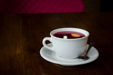 A white cup on saucer and spoon, with red tea and reflections of lamp, on dark wooden table with red chair in dark cafe