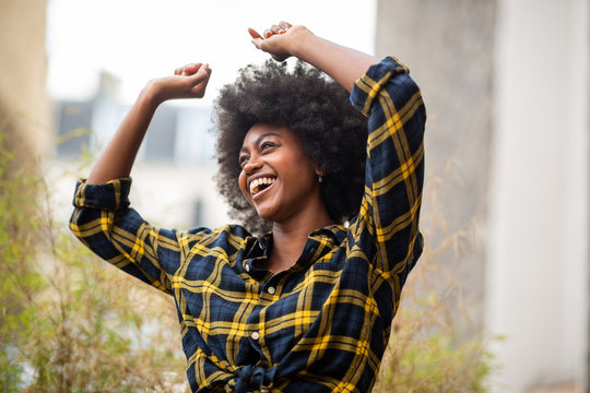 cheerful young carefree afro woman with arms raised and smiling outside