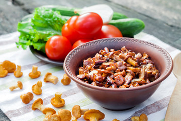Fried chanterelles with onion in rustic bowl and plate with fresh vegetables for salad on background