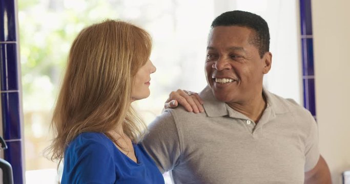 Happy older Caucasian and African American husband and wife standing in their kitchen smiling. Senior mixed race couple homeowners looking at camera. Slow motion 4k handheld