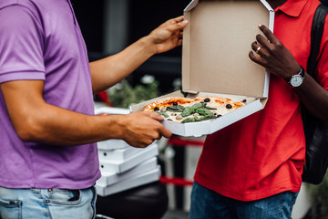 Close up photo. Courier delivery man in red uniform giving box of hot delicious pizza to customer, try it now. Candid emotion.