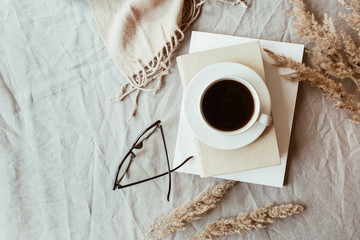 Autumn, fall composition. A cup of coffee lying on the grey linen bed with beige warm blanket, books, glasses and reeds. Lifestyle, still life concept. Flat lay, top view.