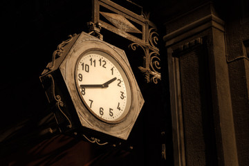 Hanging round street clock with hands, vintage and rusting, in hexagonal metal case, directly lit by strong sunlight. Monochrome image. Copy space.