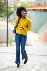 Full body happy young black woman walking with bag and mobile phone in city
