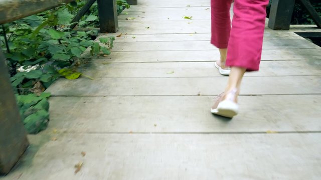 female legs in pink sneakers and burgundy trousers walk on a wooden bridge in the forest
