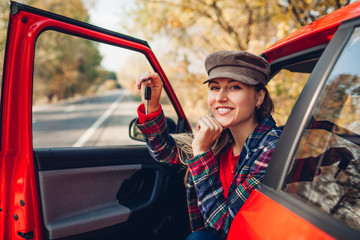 Woman holding keys of new car. Happy buyer purchased red autimobile. Driver looking at camera sitting in auto