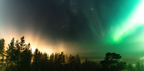Panoramic view of brilliant green Aurora shining over Swedish foggy forest landscape, light rays from a village and Northern Lights color sky in different soft colors, Northern Sweden, Scandinavia © Alexandre Patchine