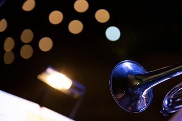 A trumpet bell with a music stand with a light in front of it in a dark stage environment and stage light bokeh in the background