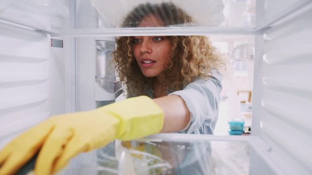 View Looking Out From Inside Empty Refrigerator As Woman Wearing Rubber Gloves Cleans Shelves