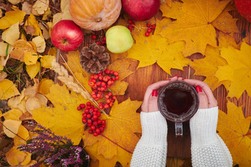 A mug of tea in a woman's hand in a sweater on the wood background with yellow falling leaves maple and cones. Hello Autumn phrase on vintage background. Hot drink. Autumn composition still life