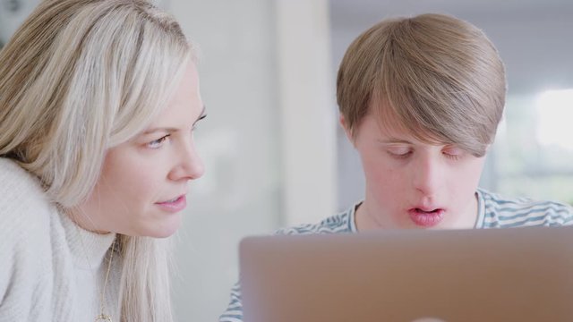 Downs Syndrome Man Sitting With Female Home Tutor Using Laptop For Lesson At Home