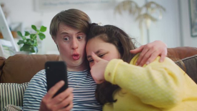 Young Downs Syndrome Couple Sitting On Sofa Using Mobile Phone To Take Selfie At Home 