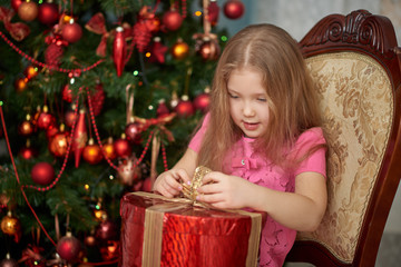 A little girl wearing light pink dress unwrapping a gift box. Decorated Christmas tree background.