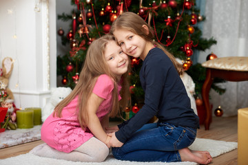 Two young sisters sitting on white carpet. Fireplace and decorated Christmas tree background.