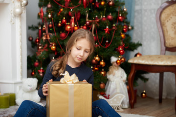 A young girl wearing a blue long sleeve shirt and jeans sitting on white carpet. Fireplace filled with toys and decorated Christmas tree background.