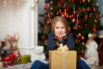 Fototapeta na wymiar A young girl wearing a blue long sleeve shirt and jeans sitting on white carpet. Fireplace filled with toys and decorated Christmas tree background.