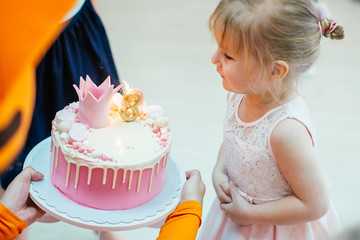 Three years old blonde adorable girl in a pink dress blowing candles on birthday pink cake with...
