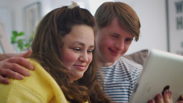 Young Downs Syndrome Couple Sitting On Sofa Watching Digital Tablet At Home