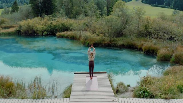 A young woman is doing yoga on her mat in the beautiful nature at a mountain lake. Outdoors meditation has fantastic health benefits. 